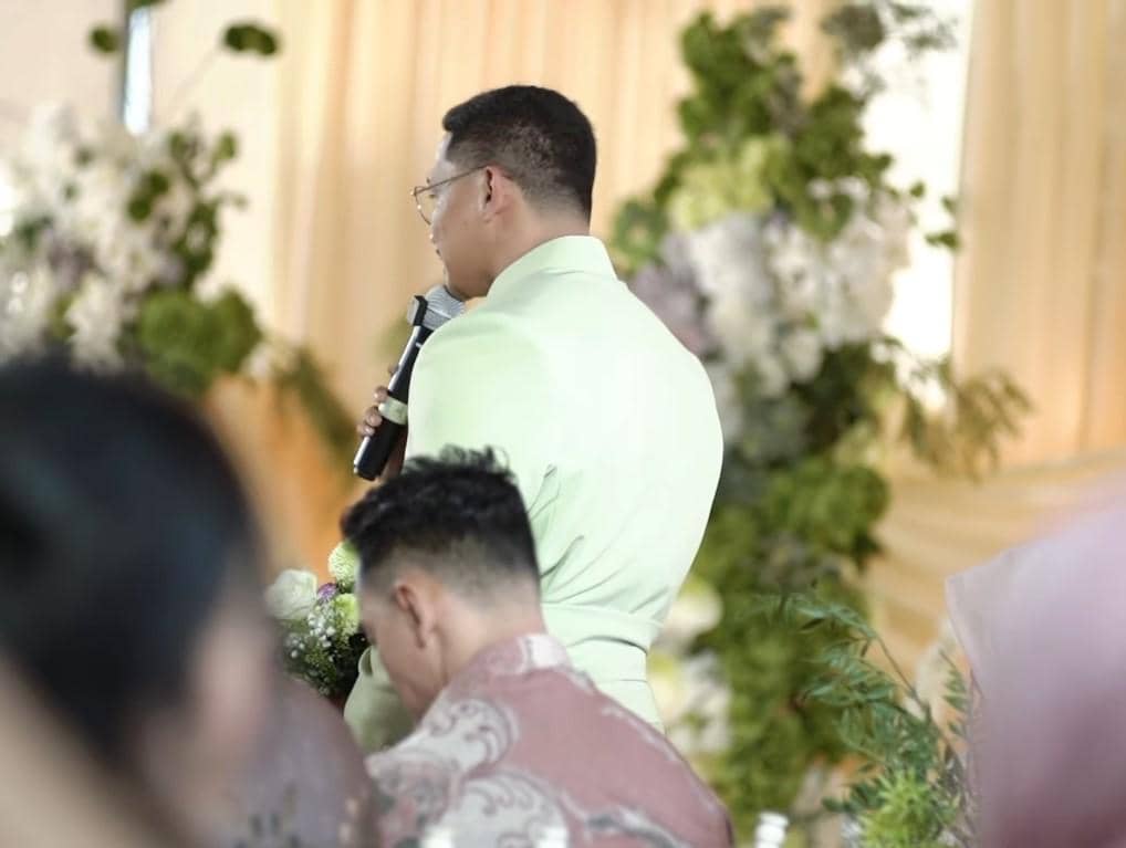 Desfian mengenakan kemeja hijau muda memegang mikrofon di acara lamaran dengan latar dekorasi bunga dan tirai krem.