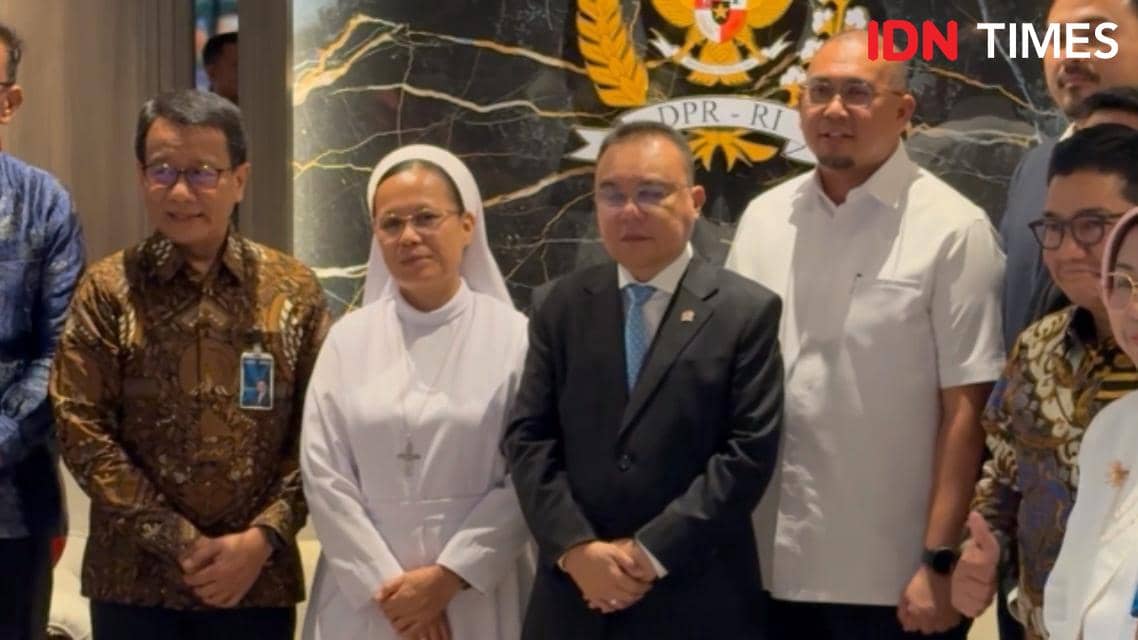 Beberapa orang berpose bersama di depan lambang DPR RI di Gedung DPR RI, termasuk pria bersetelan jas dan wanita berjubah putih.