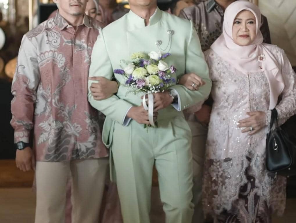 Desfian mengenakan setelan hijau muda membawa buket bunga, berjalan diapit dua orang keluarga dalam acara lamaran.