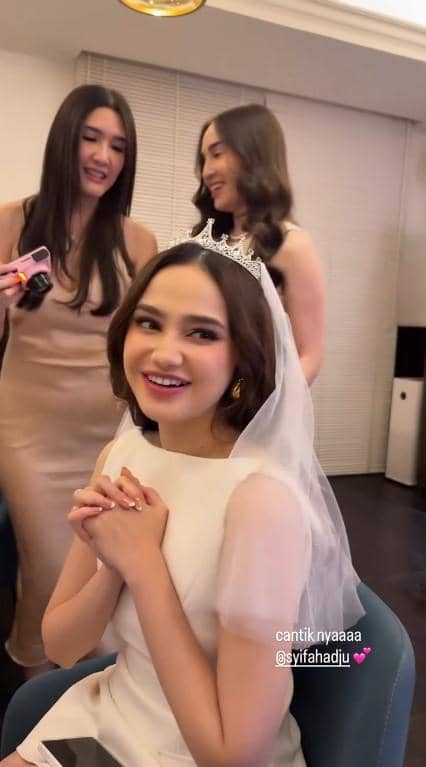 Syifa Hadju mengenakan gaun putih dan veil duduk tersenyum bahagia di acara bridal shower bersama dua temannya di dalam ruangan.