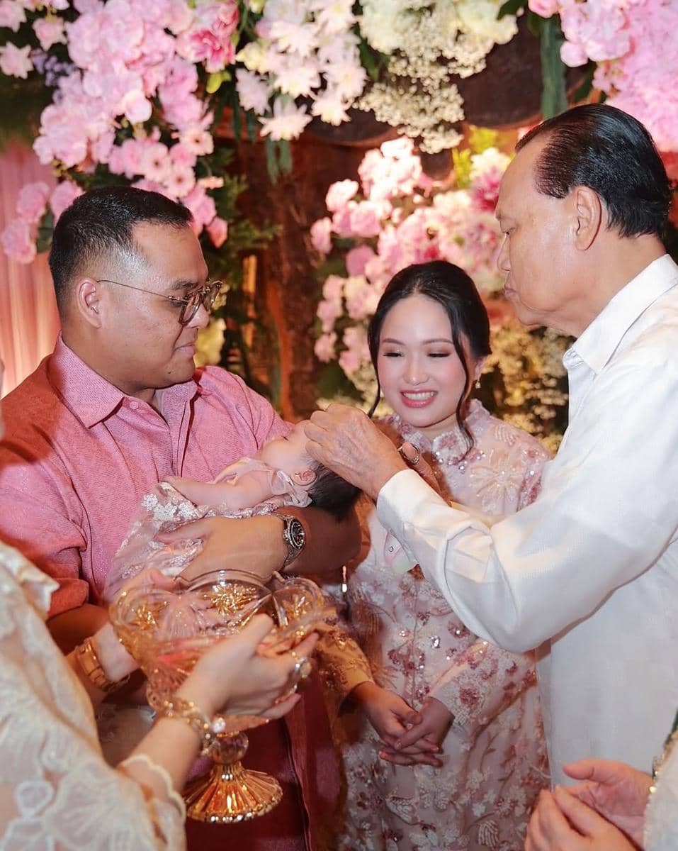 Seorang pria menggendong bayi saat prosesi akikah dengan suasana hangat, dikelilingi keluarga yang mengenakan busana bernuansa merah muda.