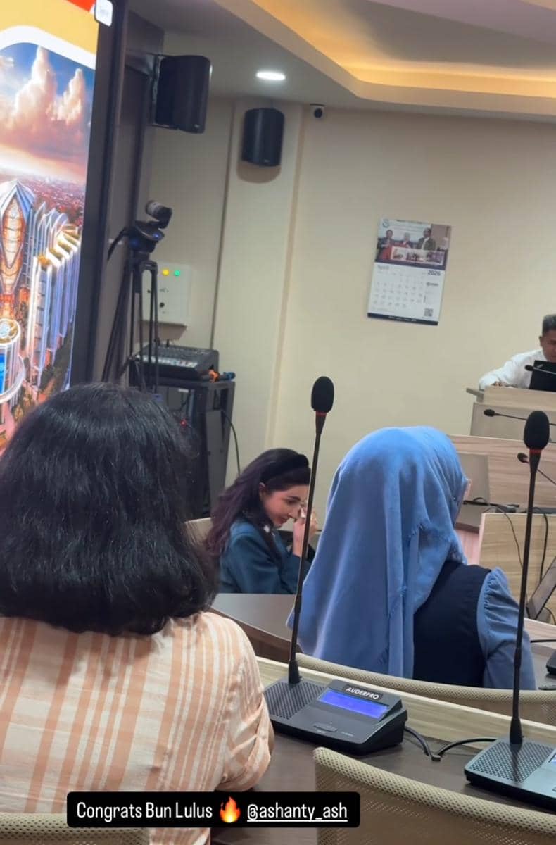 Seorang wanita duduk di ruang sidang tesis S3 di UNAIR Surabaya, dikelilingi peserta lain dan peralatan presentasi akademik.