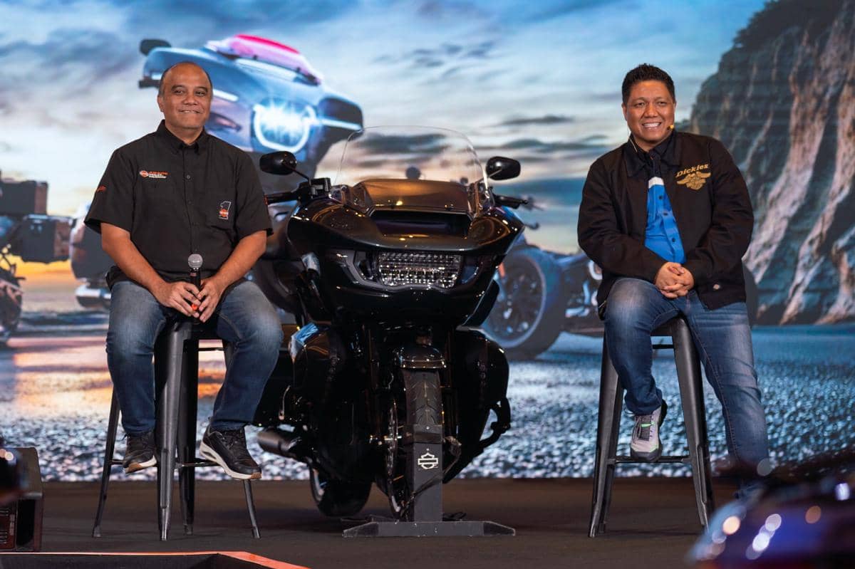 Dua pria duduk di atas panggung di samping motor besar Harley-Davidson model 2026 dengan latar belakang pemandangan jalan dan tebing.