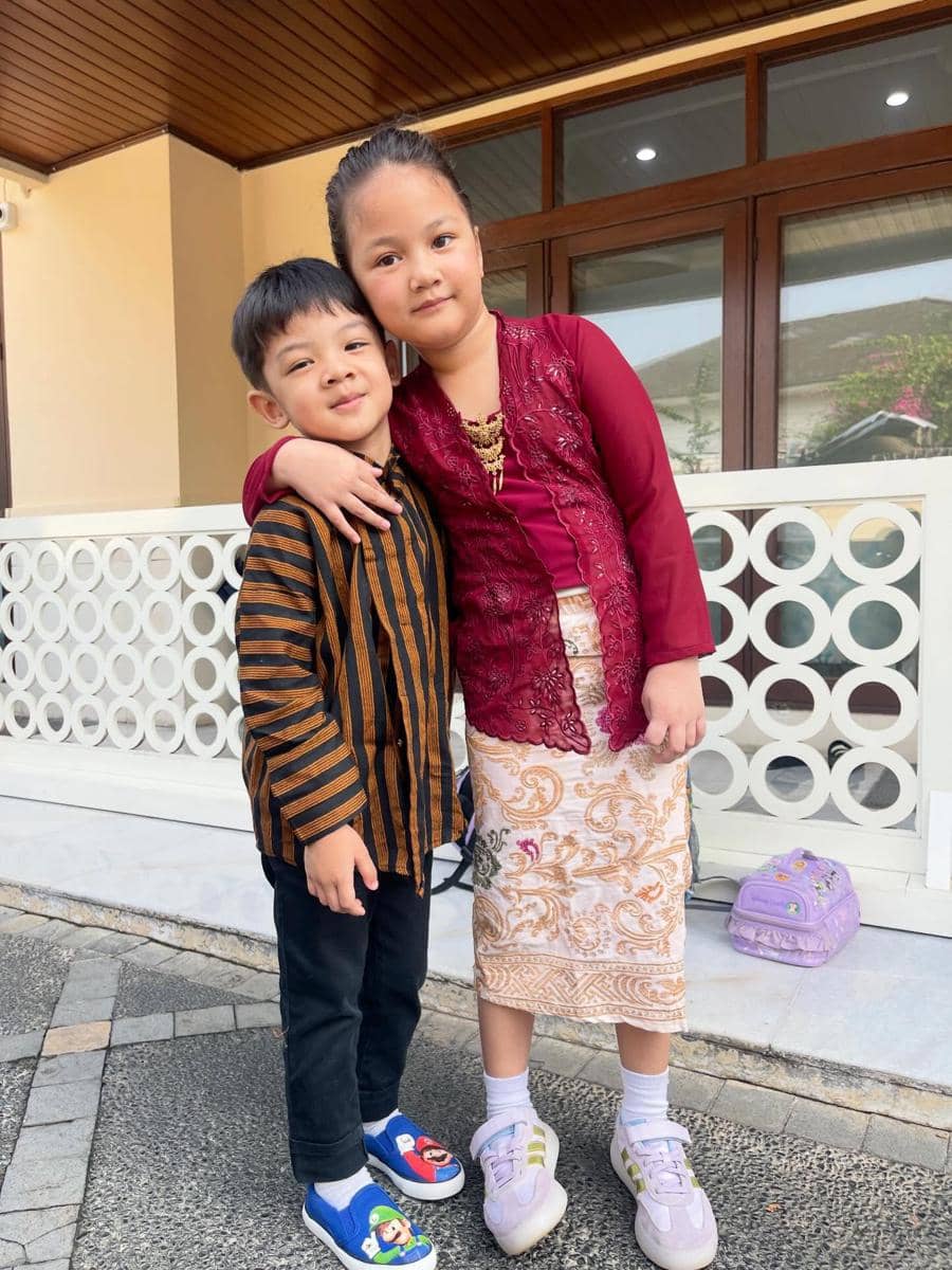 Anak Mona mengenakan pakaian tradisional, satu memakai kebaya merah dan kain batik, sementara yang lain memakai baju lurik cokelat hitam.