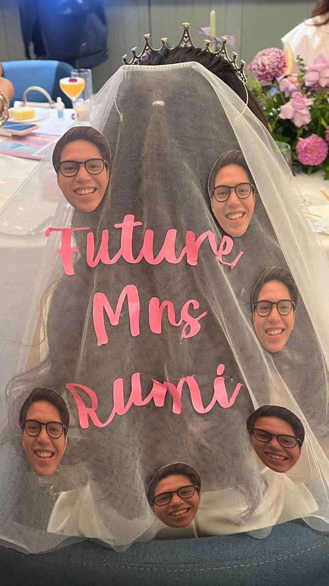 Kerudung bridal shower bertuliskan 'Future Mrs Rumi' dengan hiasan wajah pria berkacamata di atas meja dekorasi bunga.