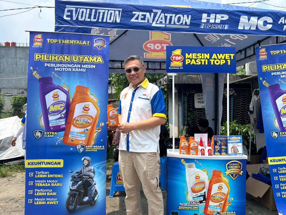 Seorang pria mengenakan kemeja putih bergaris biru dan kuning memegang botol oli motor TOP 1 di stan promosi luar ruangan.