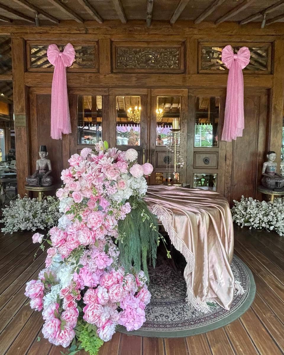 Dekorasi akikah dengan nuansa pink dan emas, dihiasi bunga mawar dan peoni di atas meja berlapis kain satin di ruangan kayu tradisional.