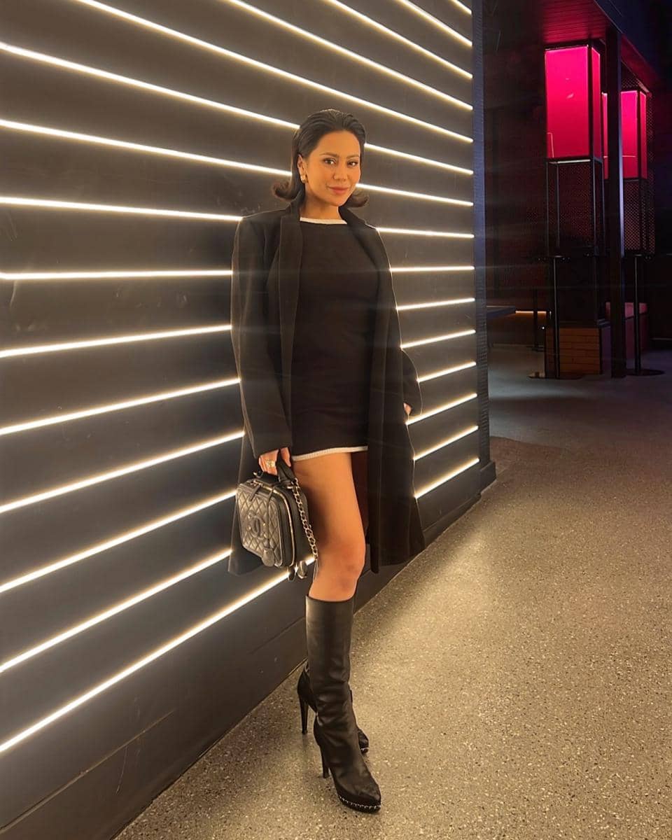 Vicky berpose di depan dinding dengan lampu garis horizontal, mengenakan gaun hitam pendek, mantel panjang, dan sepatu bot tinggi hitam.