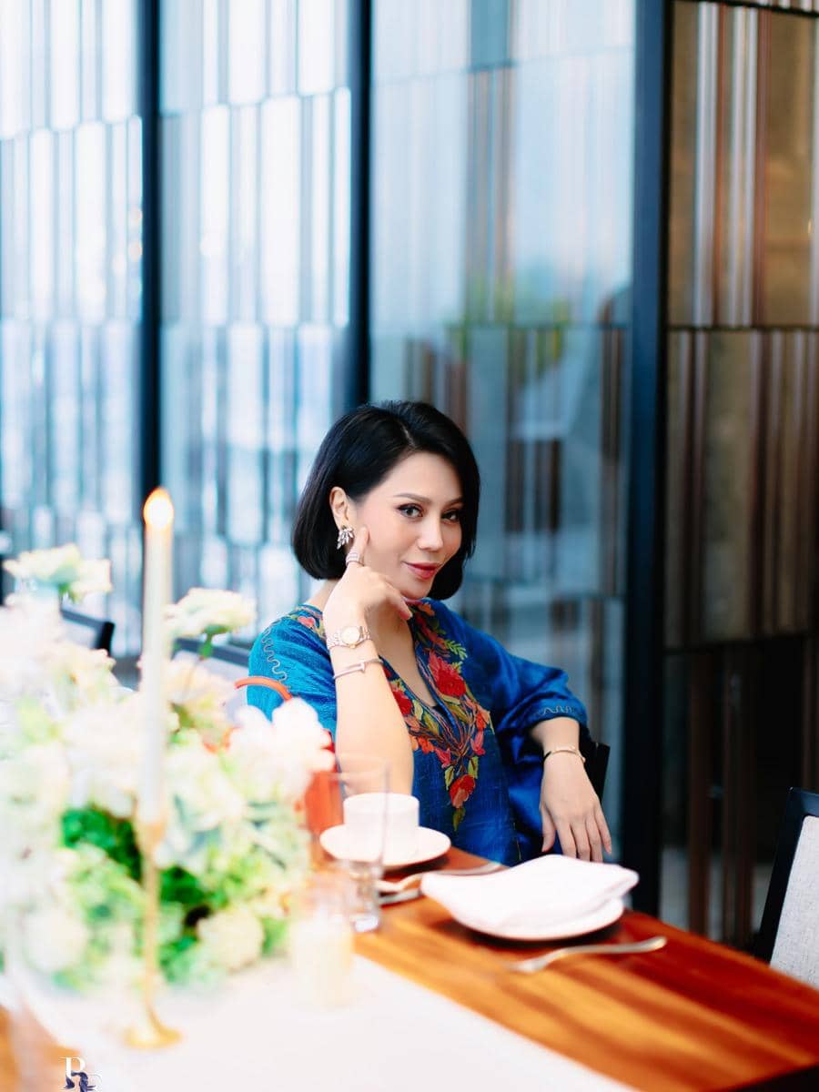 Vicky Zainal berambut pendek mengenakan pakaian biru bermotif duduk di meja makan elegan dengan dekorasi bunga dan lilin.