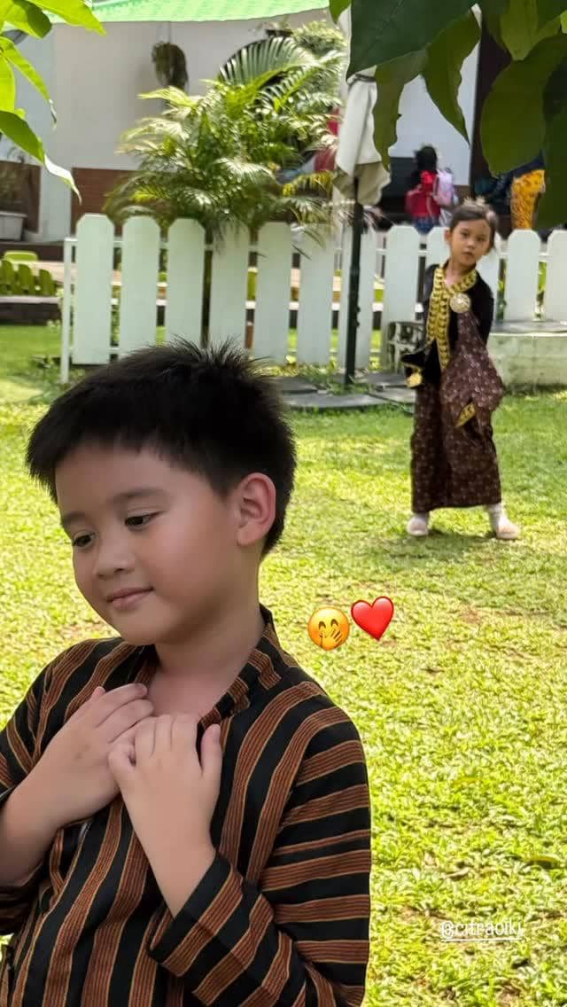 Anak Citra Kirana mengenakan pakaian tradisional Jawa berpose di taman, dengan seorang anak perempuan di latar belakang juga memakai kebaya.