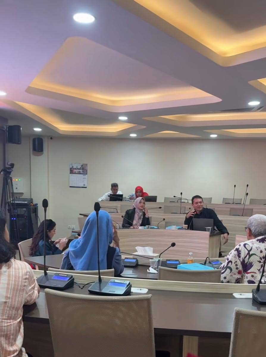 Seorang wanita sedang menjalani sidang tesis S3 di ruang pertemuan universitas dengan beberapa dosen dan peserta lainnya di Surabaya.