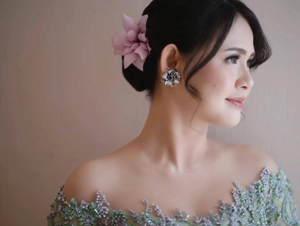 Maizura mengenakan kebaya hijau berhias payet dengan tatanan rambut sanggul dan hiasan bunga ungu muda di sisi kepala.