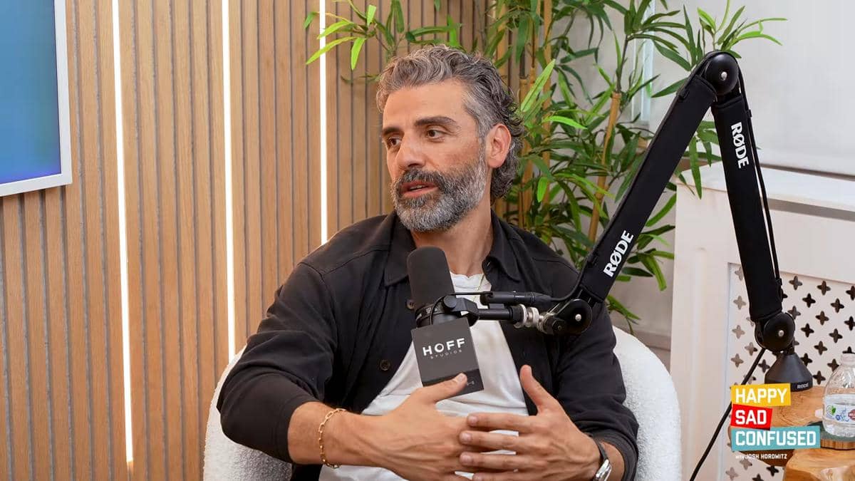 Oscar Isaac berbicara di depan mikrofon saat menjadi tamu di podcast Happy Sad Confused dengan latar ruangan kayu dan tanaman hias.