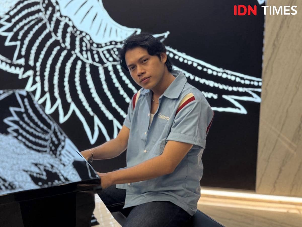 Seorang pria mengenakan kemeja biru duduk di depan piano hitam dengan latar belakang bergambar sayap berwarna putih.