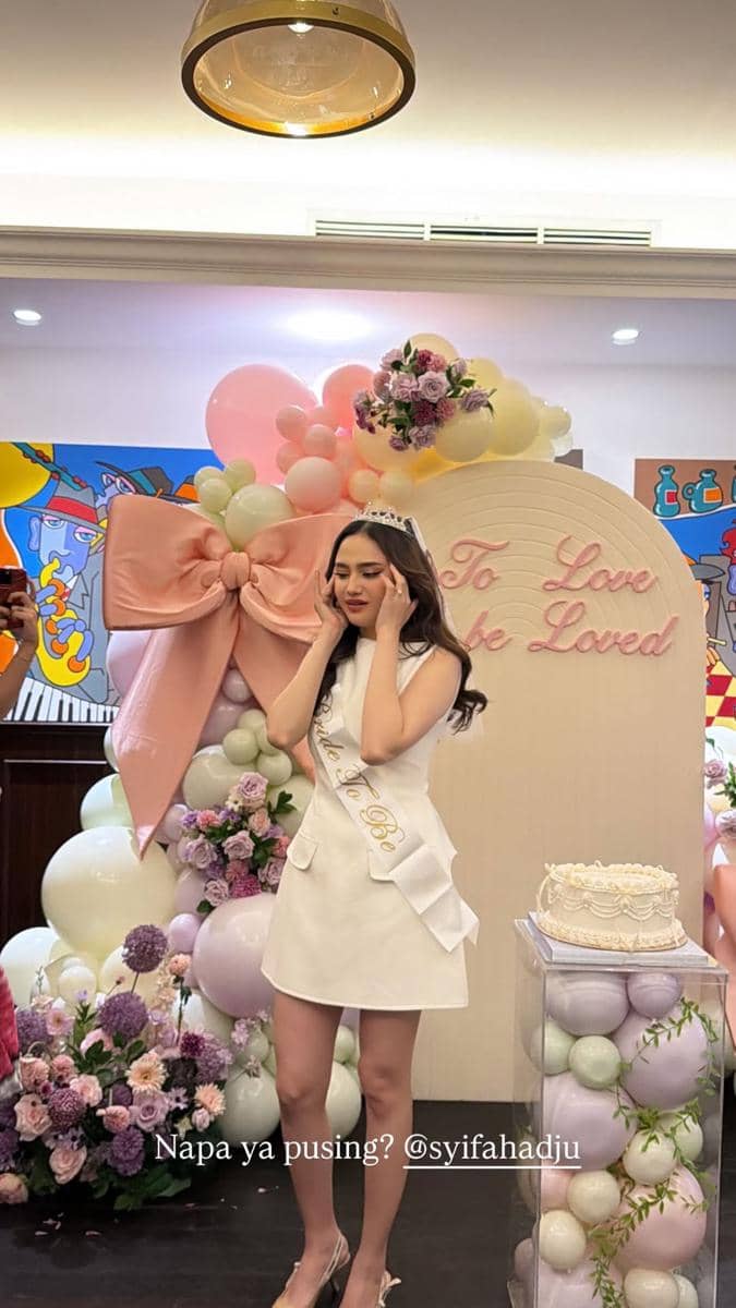 Syifa Hadju mengenakan gaun putih dan selempang bride to be berdiri di depan dekorasi balon pastel dan bunga saat acara bridal shower.