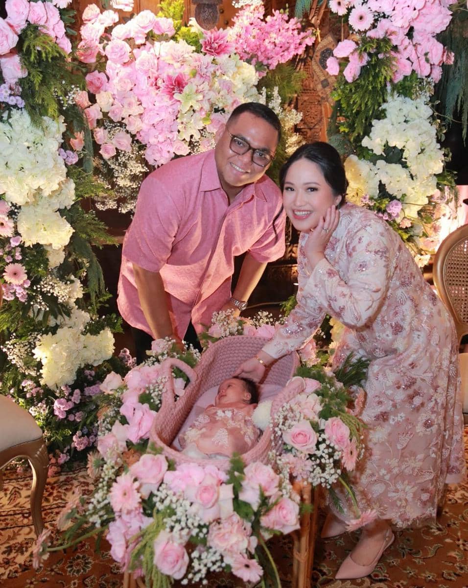 Pasangan tersenyum berdiri di samping bayi yang terbaring di keranjang berhias bunga warna pink dan putih dalam acara akikah.