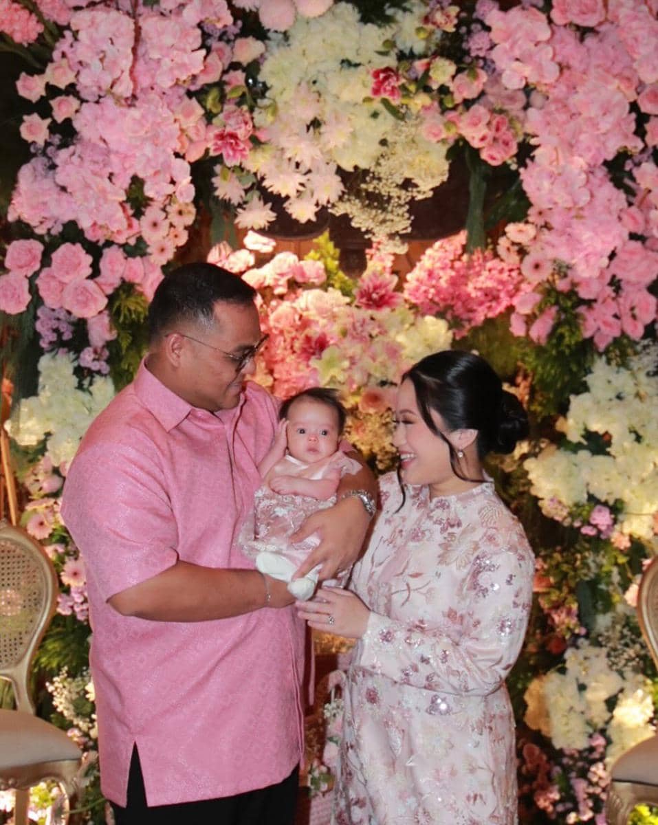 Pasangan suami istri mengenakan busana bernuansa pink sambil menggendong bayi di depan dekorasi bunga warna merah muda dan putih.