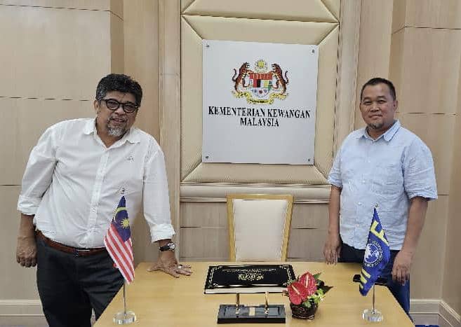 Dua pria berdiri di ruang pertemuan dengan latar belakang logo Kementerian Keuangan Malaysia dan meja berhiaskan bendera serta bunga.