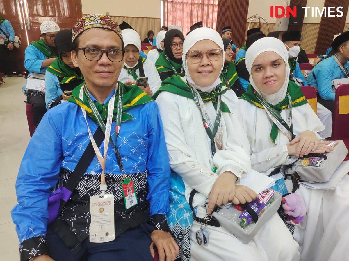 Tiga calon jemaah haji mengenakan seragam biru dan putih dengan syal hijau duduk bersama di ruang tunggu keberangkatan haji.