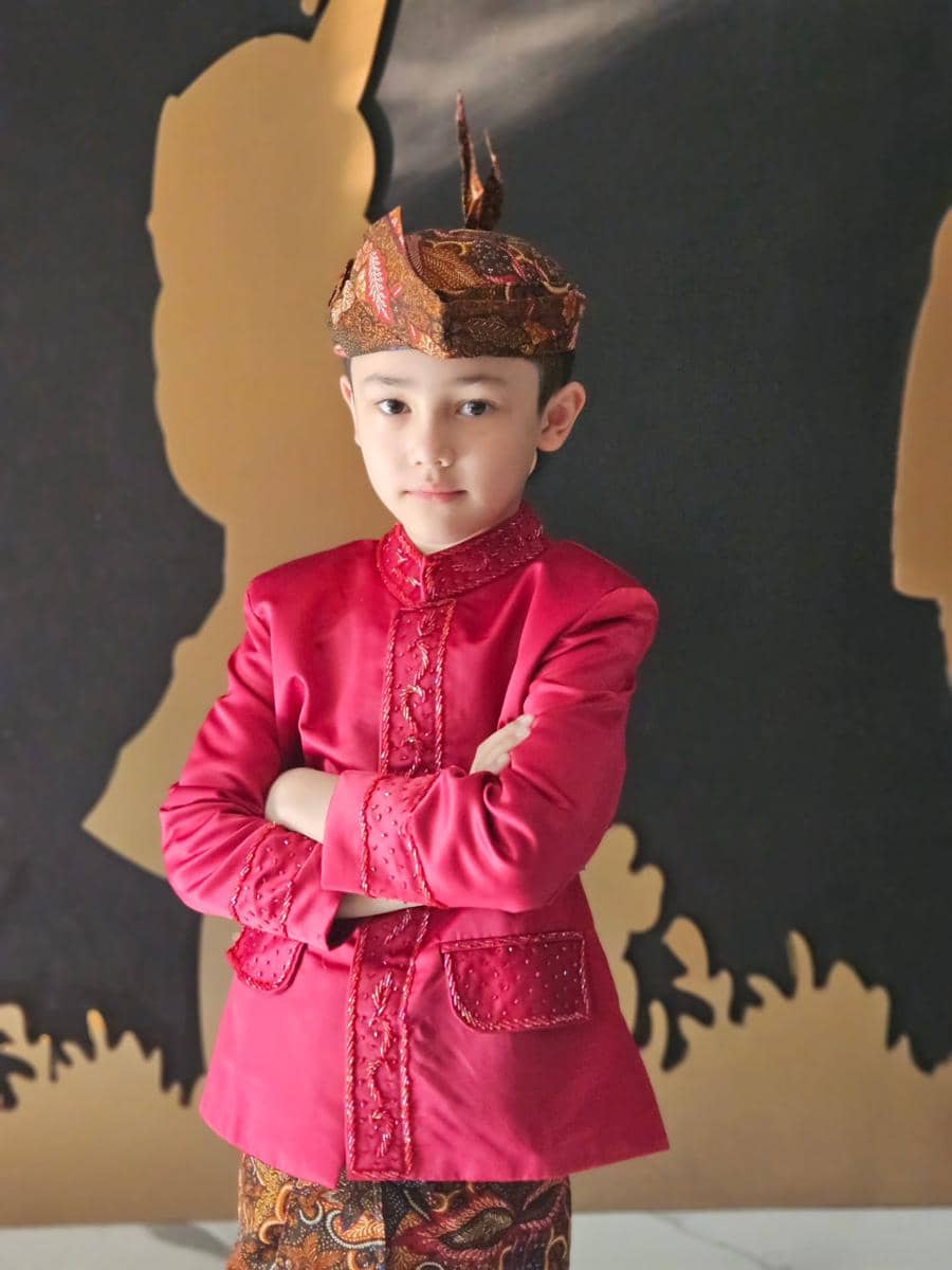 Anak Andhika Pratama mengenakan pakaian adat berwarna merah dan batik, berpose dengan tangan terlipat di depan latar dekoratif Hari Kartini.