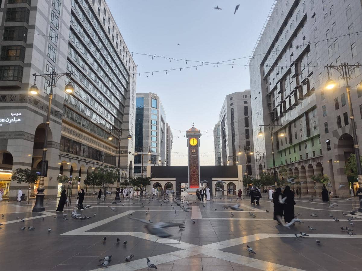 Deretan gedung hotel tinggi di sekitar Masjid Nabawi di Madinah dengan pejalan kaki dan burung merpati di area plaza.