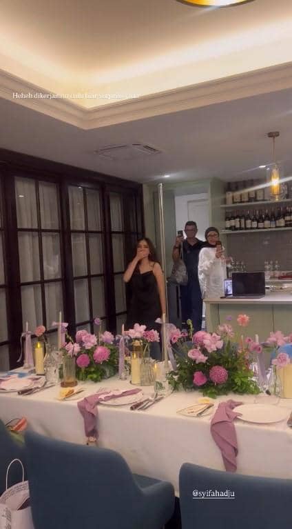 Syifa Hadju mengenakan gaun hitam berdiri di ruang makan yang dihiasi bunga ungu dan merah muda untuk acara bridal shower.