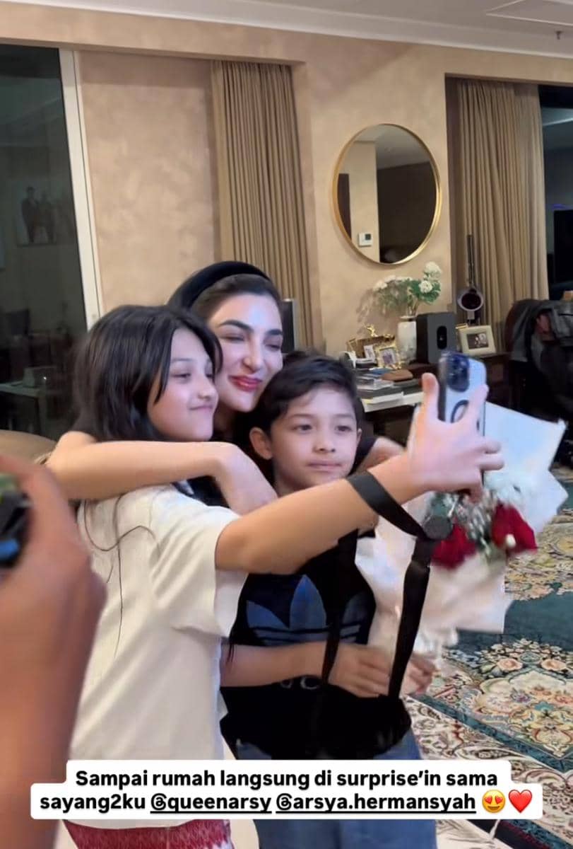 Seorang wanita berpose sambil berpelukan dengan dua anak yang sedang mengambil swafoto di ruang tamu rumah yang hangat dan rapi.