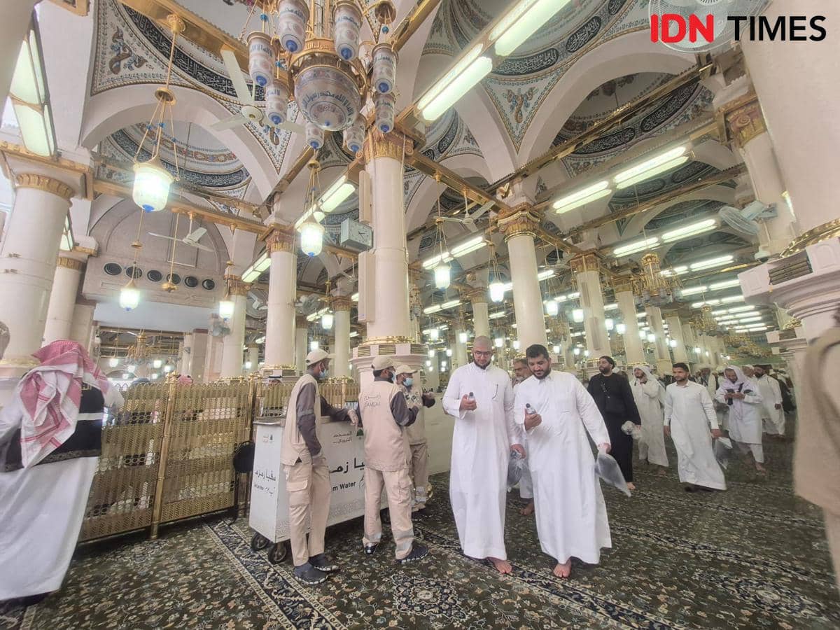 Jemaah berpakaian gamis dan petugas masjid berjalan di dalam Masjid Nabawi dengan interior megah berhiaskan lampu gantung dan pilar putih.