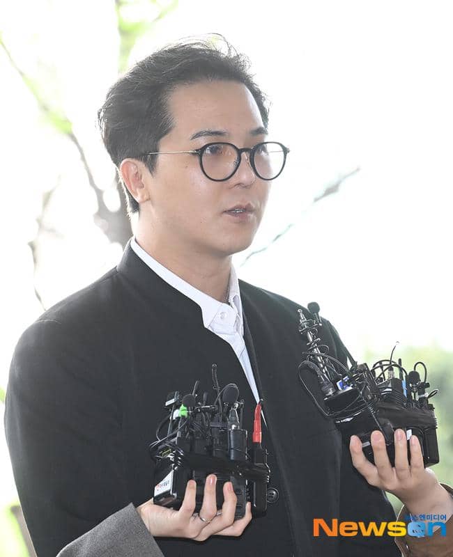 Seorang pria mengenakan kacamata dan jas hitam memegang beberapa mikrofon di luar ruangan dengan latar belakang terang.