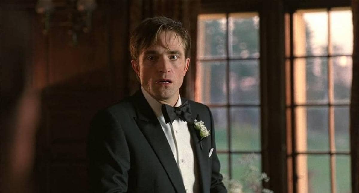 Seorang pria mengenakan setelan jas hitam dengan dasi kupu-kupu dan bunga di dada berdiri di dalam ruangan dengan jendela besar di belakangnya.