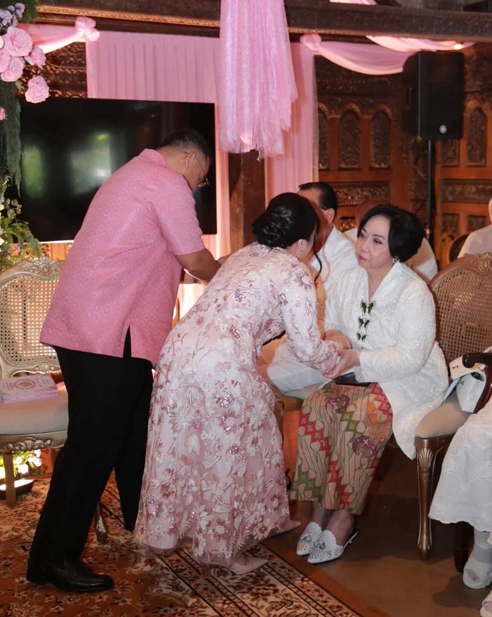 Seorang wanita mengenakan kebaya pink bersalaman dengan tamu yang duduk mengenakan kebaya putih dalam acara akikah bernuansa tradisional.