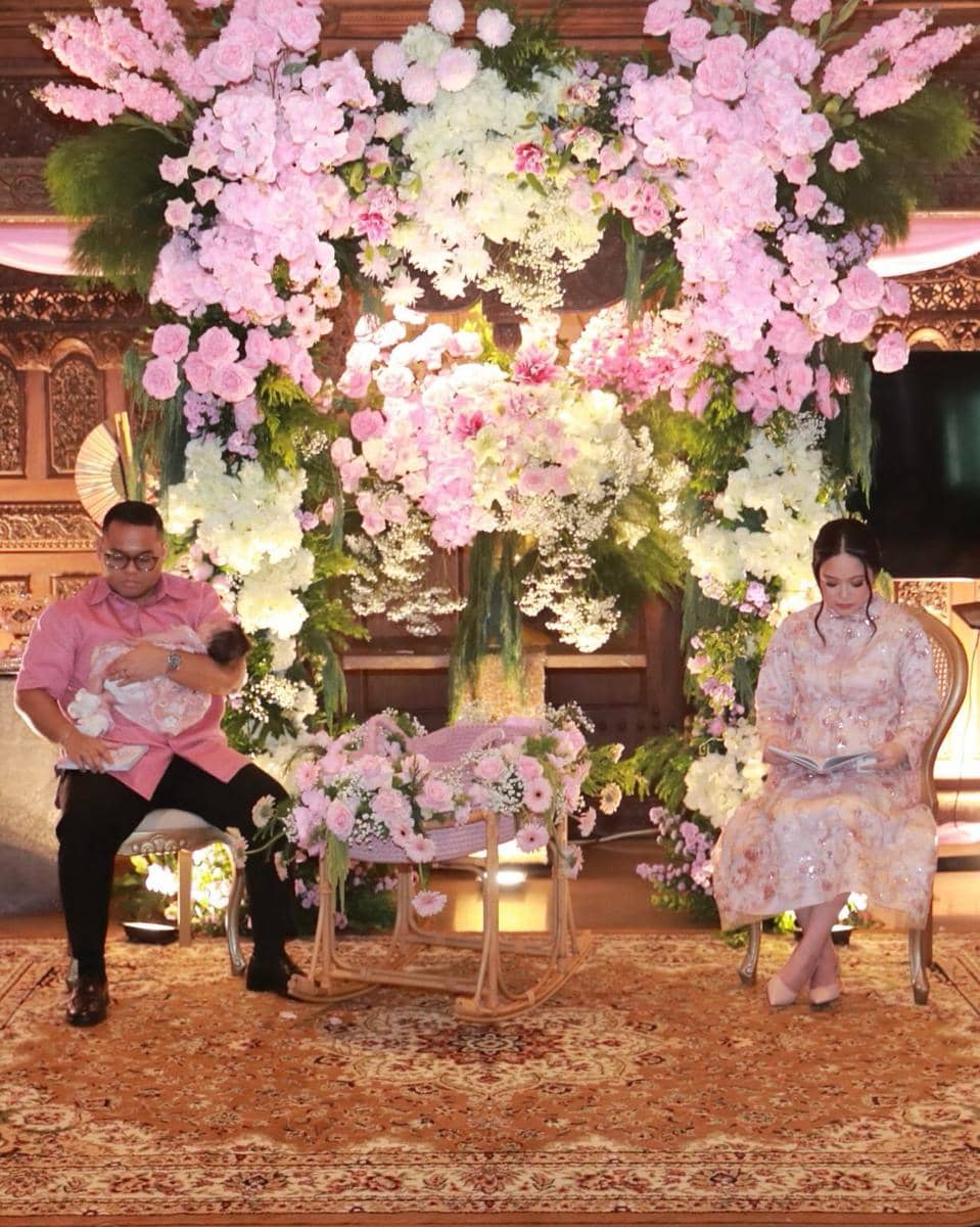 Pasangan duduk di depan dekorasi akikah bernuansa bunga pink dan putih dengan keranjang bayi berhias bunga di tengah ruangan.