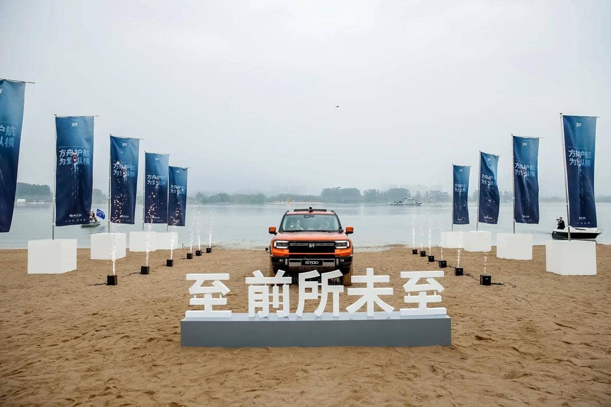 SUV Jetour G700 berwarna oranye dipamerkan di tepi pantai dengan latar belakang spanduk biru dan air mancur kecil di sekitarnya.