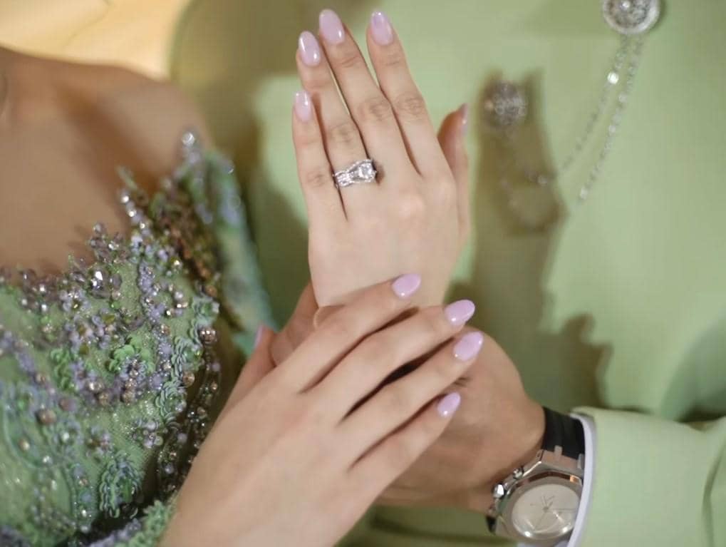 Tangan pasangan mengenakan cincin lamaran dengan busana berwarna hijau pastel berhias payet dan jam tangan elegan.