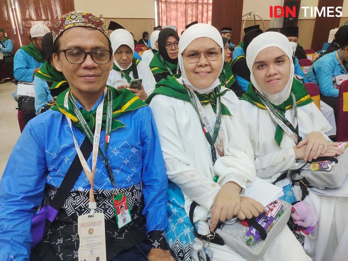 Tiga calon jemaah haji mengenakan pakaian seragam haji berwarna putih dan biru dengan syal hijau duduk bersama di ruang keberangkatan.
