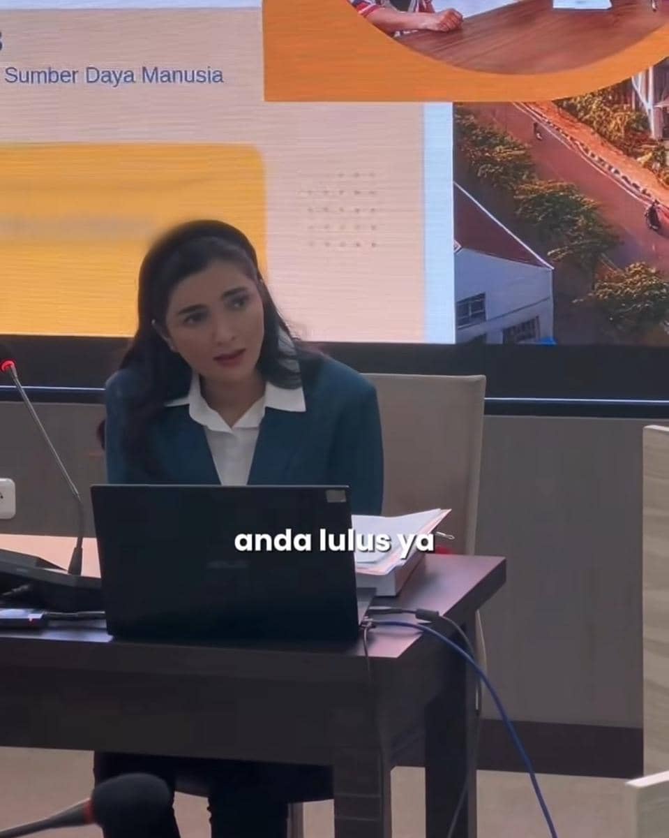 Seorang wanita mengenakan jas biru dan kemeja putih sedang duduk di depan laptop saat menjalani sidang tesis di UNAIR Surabaya.