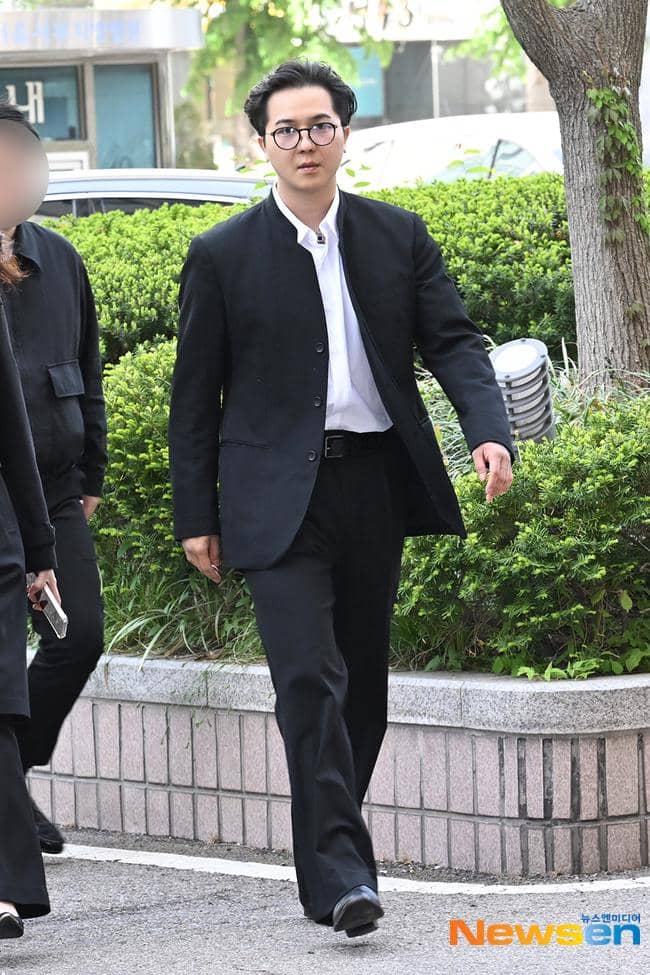 Seorang pria mengenakan setelan jas hitam dan kemeja putih berjalan di area luar gedung dengan latar pepohonan hijau.