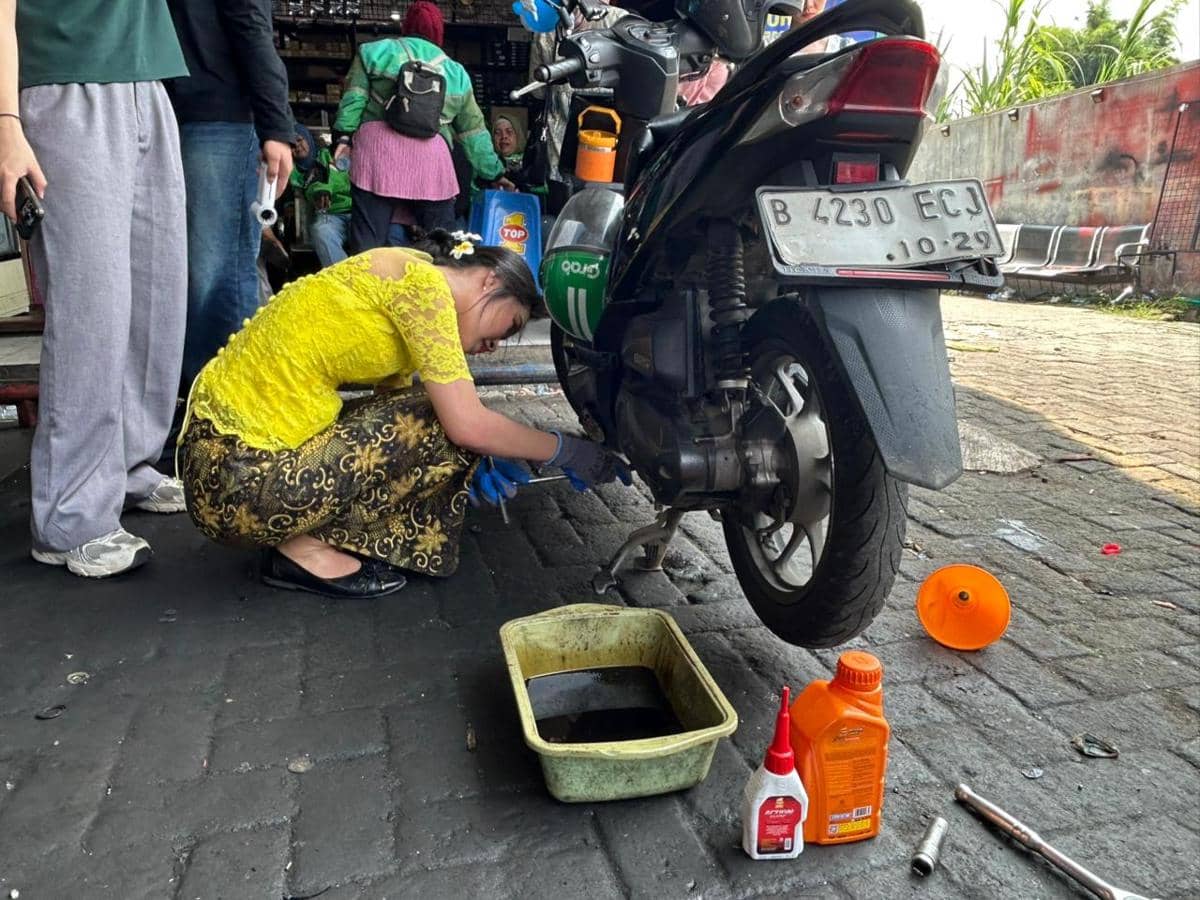 Seorang driver ojek online wanita mengenakan kebaya kuning sedang mengganti oli motor di bengkel dengan peralatan di sekitarnya.