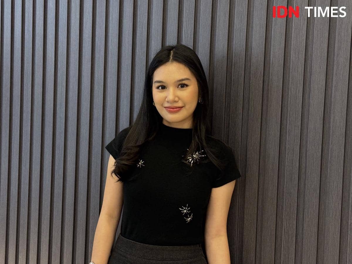 Seorang perempuan berambut panjang mengenakan kaus hitam berhias bros berdiri di depan dinding bertekstur garis vertikal.