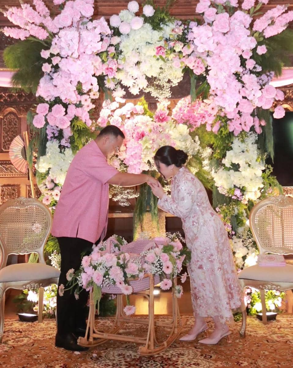 Seorang wanita mengenakan kebaya berwarna pink muda mencium tangan pria berbaju pink di acara akikah dengan dekorasi bunga warna pastel.