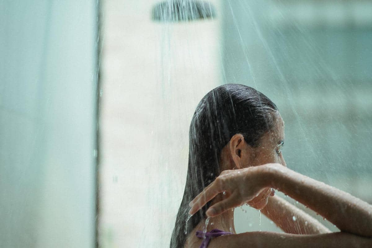 Seorang perempuan sedang mandi di bawah pancuran air dingin dengan rambut basah dan suasana kamar mandi yang lembap.