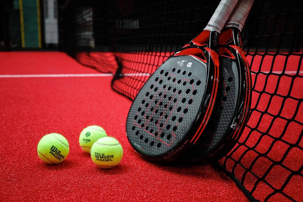 Dua raket padel berwarna hitam dan merah bersandar di jaring lapangan dengan tiga bola padel kuning di lantai merah.