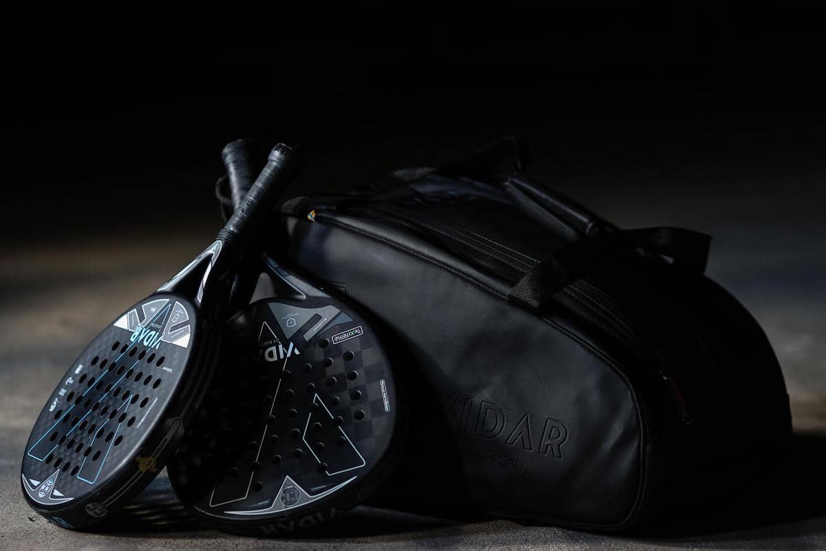 Dua raket padel berwarna hitam bersandar pada tas olahraga hitam di permukaan gelap dengan pencahayaan redup.