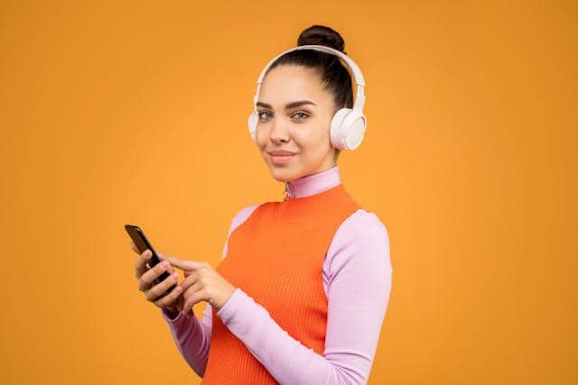 Seorang wanita mengenakan headphone putih sambil memegang ponsel dan tersenyum dengan latar belakang oranye cerah.