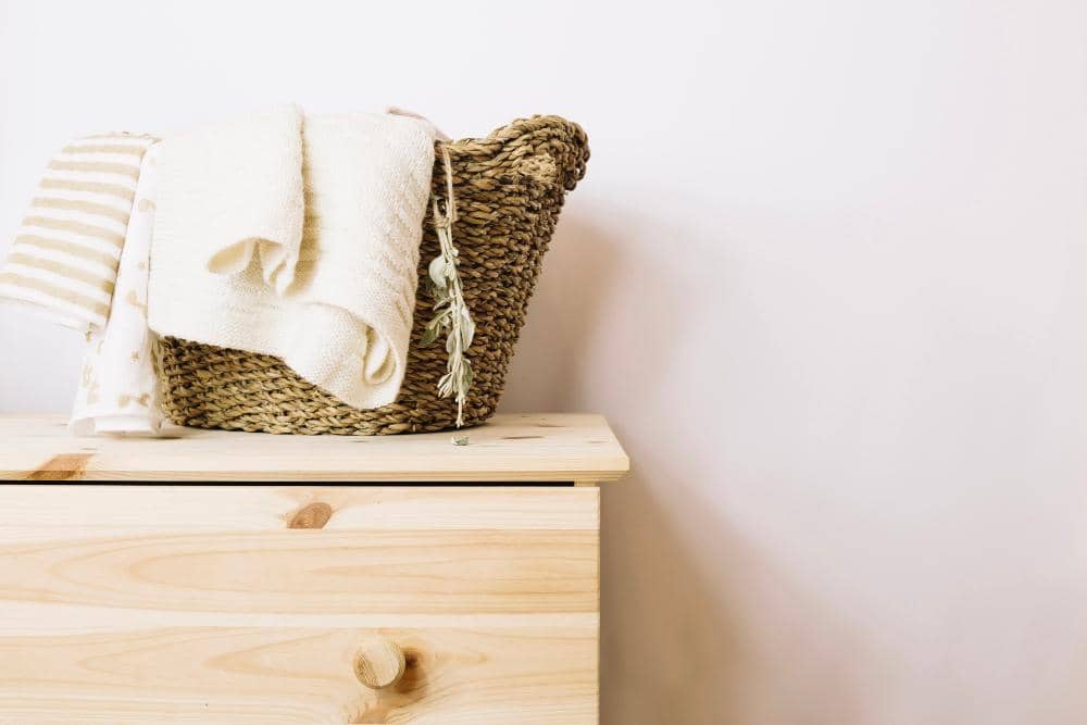 Keranjang anyaman berisi handuk putih dan kain bergaris diletakkan di atas meja kayu terang dengan latar dinding putih.
