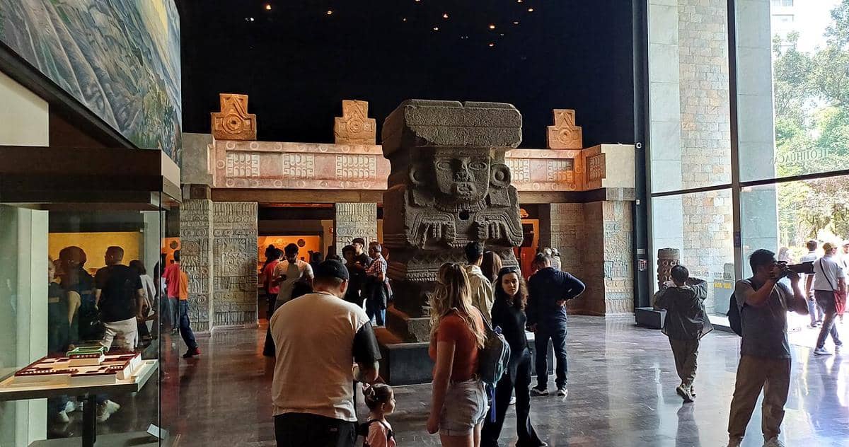 Pengunjung melihat patung batu besar bergaya pra-Columbus di dalam Museum Nasional Antropologi dengan dinding berpola dan pencahayaan alami.