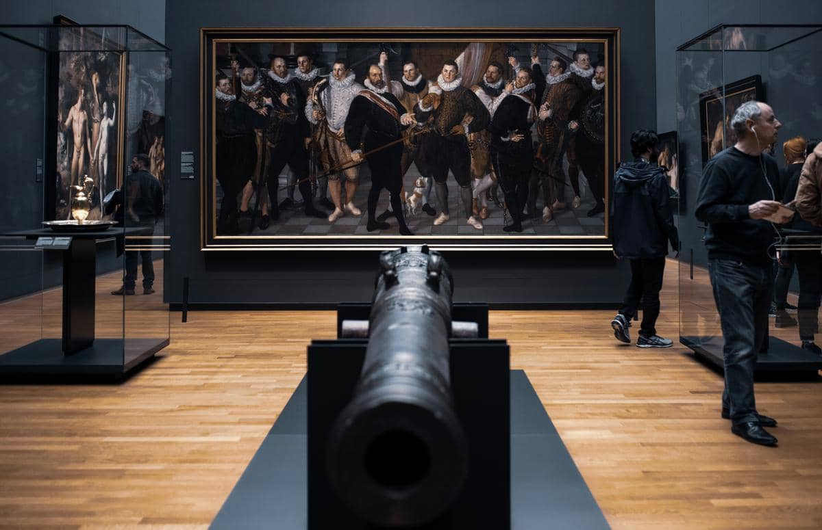 Interior Rijksmuseum dengan lukisan besar bergaya klasik, meriam kuno di tengah ruangan, dan pengunjung yang melihat karya seni.