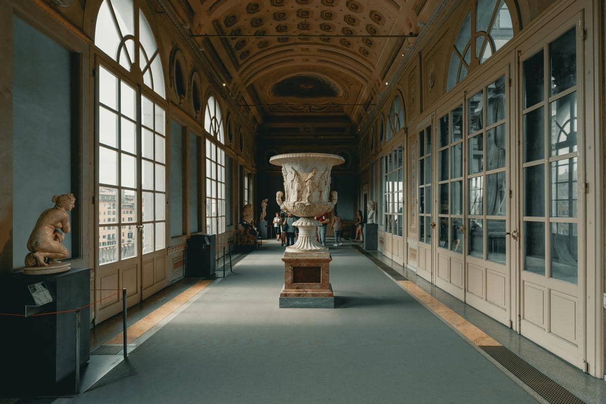 Lorong dalam Galeri Uffizi dengan patung marmer besar di tengah ruangan, dikelilingi jendela tinggi dan langit-langit berhias ukiran klasik.