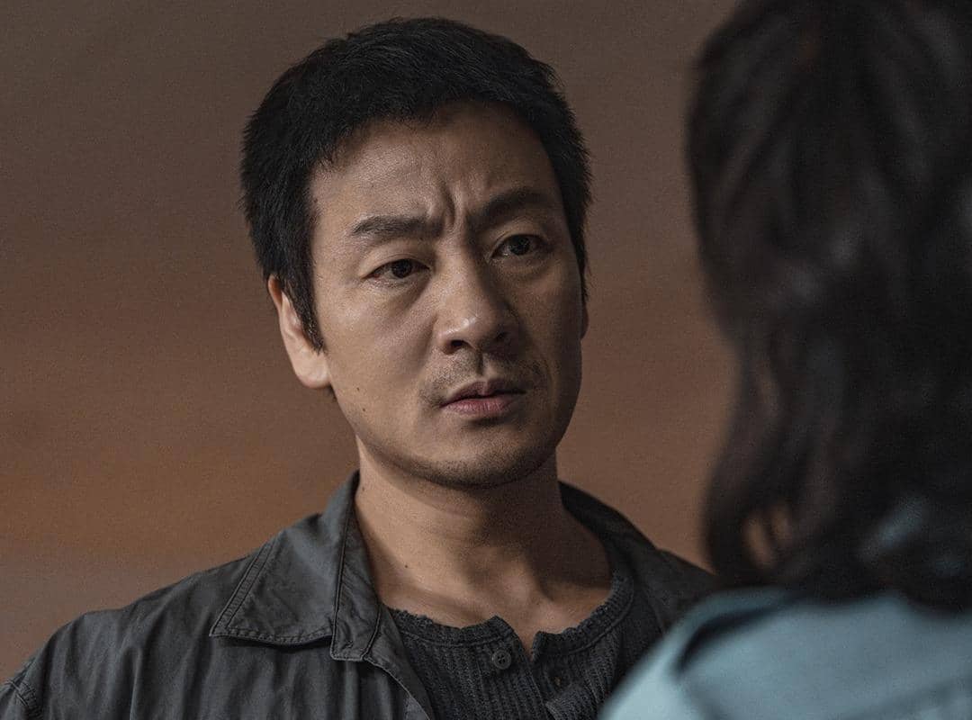 Seorang pria berwajah serius mengenakan jaket abu-abu sedang menatap seseorang di depannya dalam adegan drama Korea The Scarecrow.