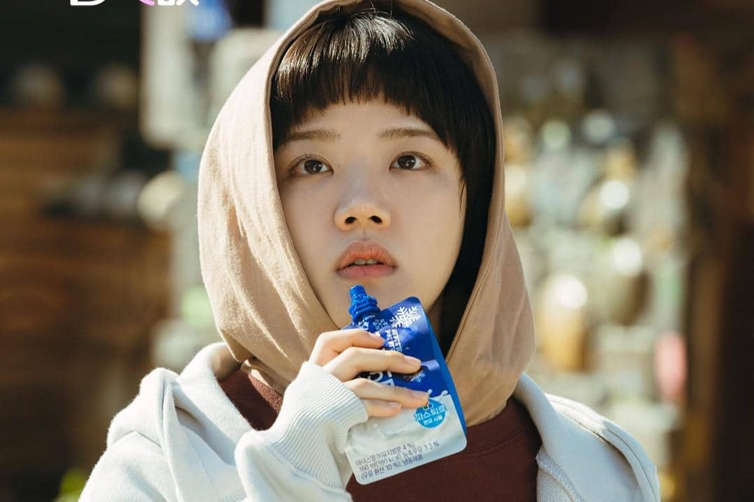 Seorang perempuan berjaket krem dengan tudung di kepala memegang minuman kemasan biru dalam adegan drama Absolute Value of Romance.